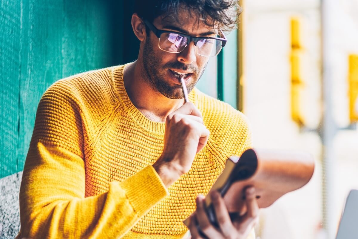 Shutterstock: Concentrated young man in eyeglasses thinking on writing essay in notepad at modern laptop computer connected to wireless high speed internet.Clever student thinking on checklist sitting in coworking
