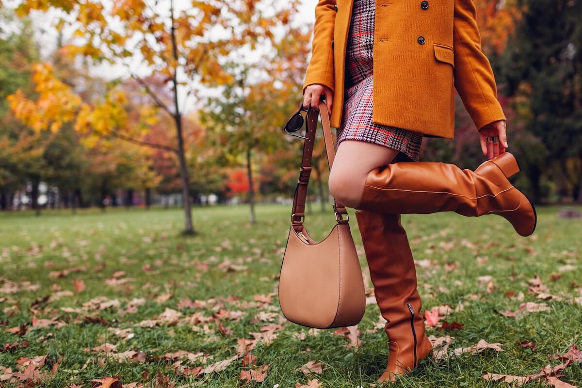 Shutterstock: Orange knee high boots. Fashionable woman wearing stylish blazer plaid mini skirt walking in fall park with beige handbag among leaves. Copy space