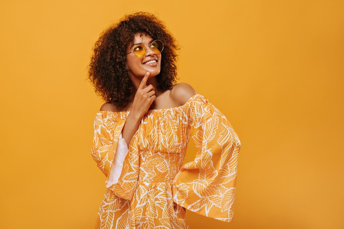 Shutterstock: Dark-skinned woman in sunglasses smiling on isolated backdrop. Pensive girl in summer clothes posing on yellow background..