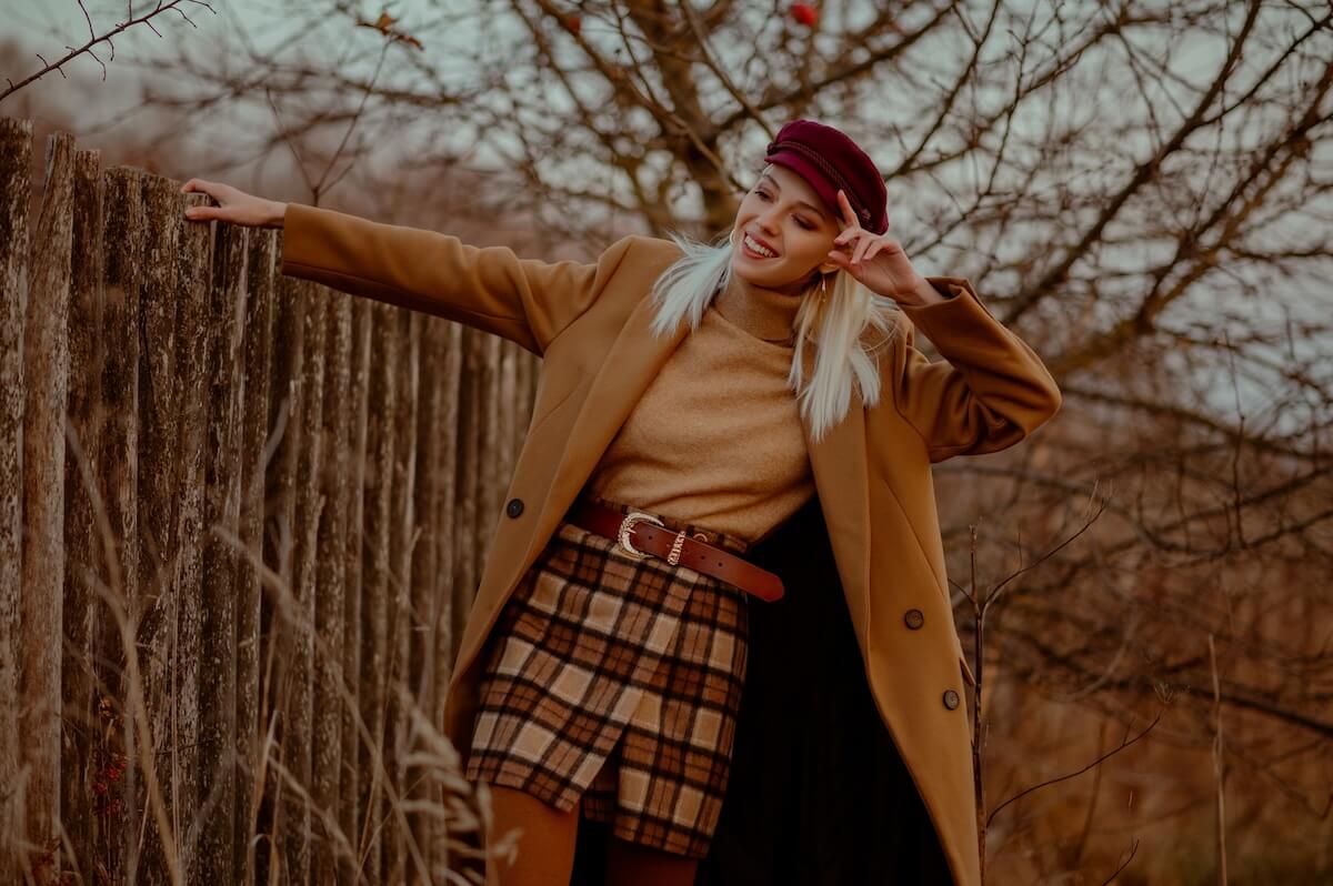 Shutterstock: Happy smiling fashionable blonde woman wearing trendy autumn outfit with classic brown coat, trendy cap, beige turtleneck sweater, leather belt, plaid mini skirt, posing outdoor. Copy, empty space