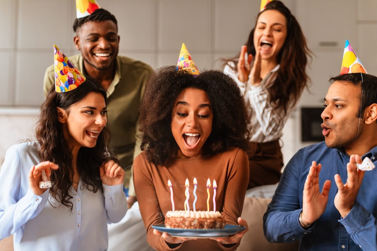 Shutterstock: Multiracial friends surround a woman holding a birthday cake, blowing out candles in a moment of excitement together