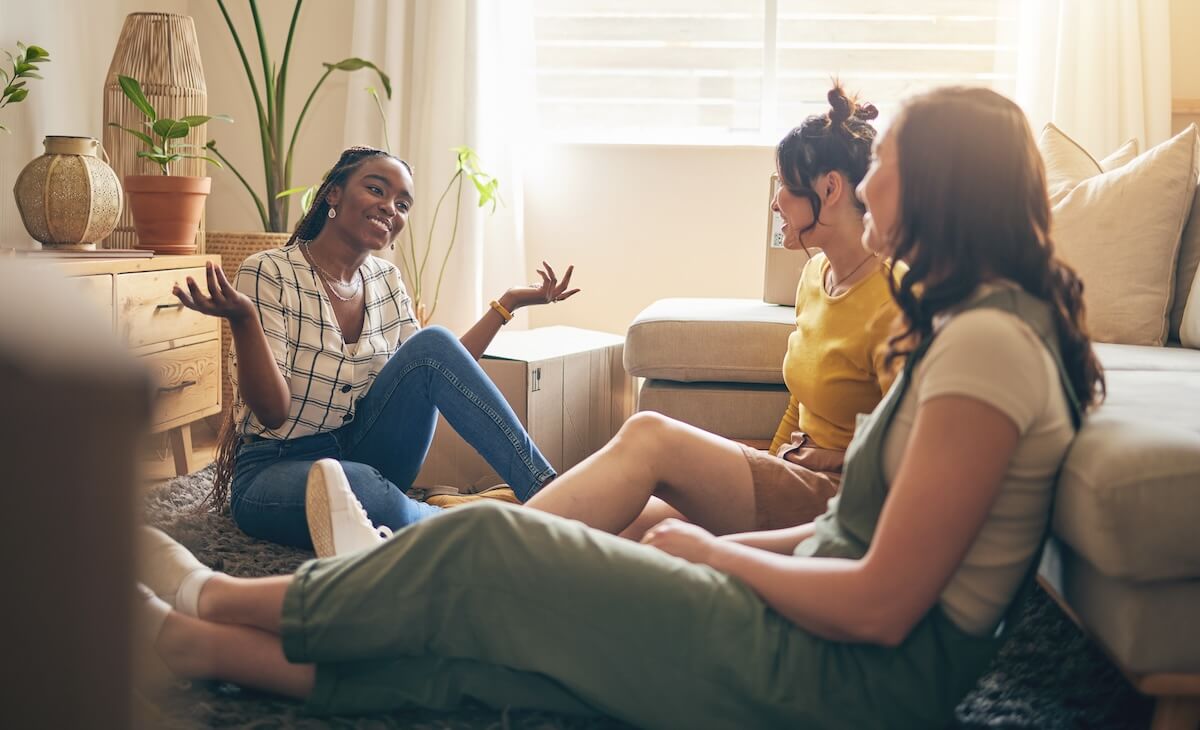 Shutterstock: Happy, gossip and friends on a floor relax, talking and bond with advice in house together. Conversation, drama and women with diversity in a living room speaking, chilling and enjoy weekend freedom
