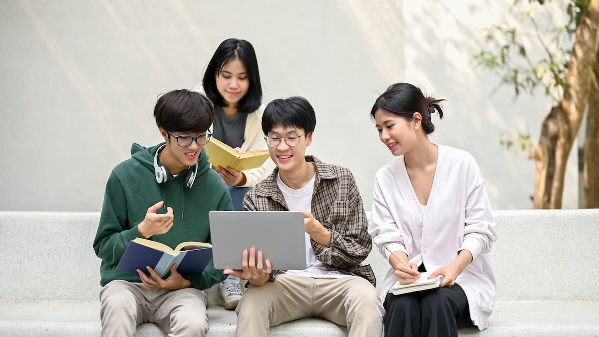 Shutterstock: Group of happy young Asian college students sitting on a bench, looking at a laptop screen, discussing and brainstorming on their school project together.