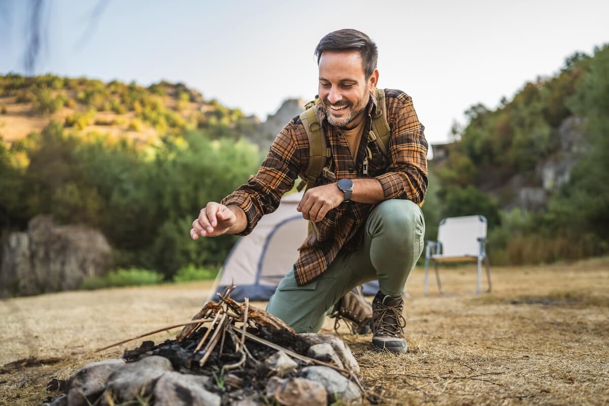 Shutterstock: A man wearing a backpack and flannel shirt is set up a campfire with sticks, while a tent and natural surroundings are visible in the background at an outdoor campsite