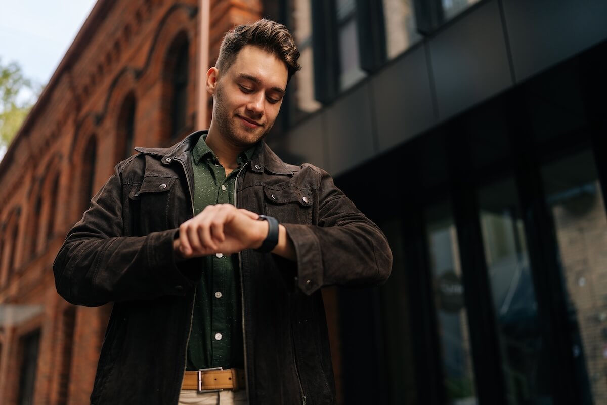 Shutterstock :Confident successful businessman wearing brown suede jacket and green shirt smiling while checking time on smartwatch standing by office building, exuding punctuality and assurance.