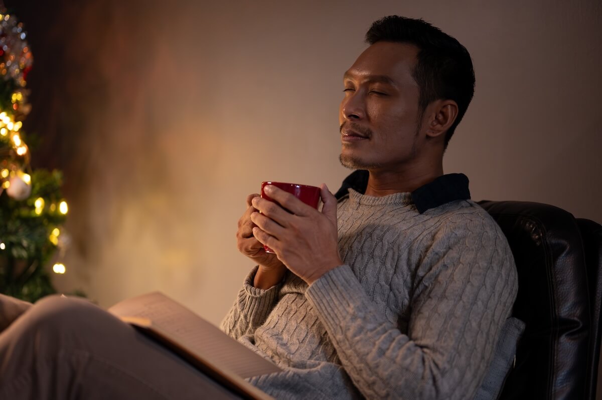 Shutterstock: Side view image of a relaxed and calm Asian man in a cosy sweater, eyes closed, smelling his coffee, enjoying his coffee or hot cocoa in a living room on his Christmas night at home alone.