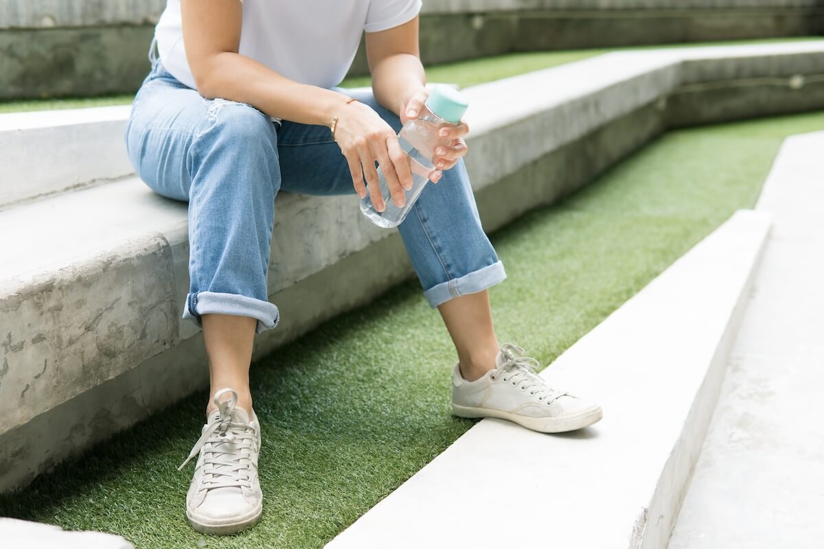 Shutterstock: Portrait of beautiful & stylish young woman, wearing white shirt, jeans and sneakers, sit on modern outdoor cement stair and hold personal resusable water bottle. Stop single use plastic concept