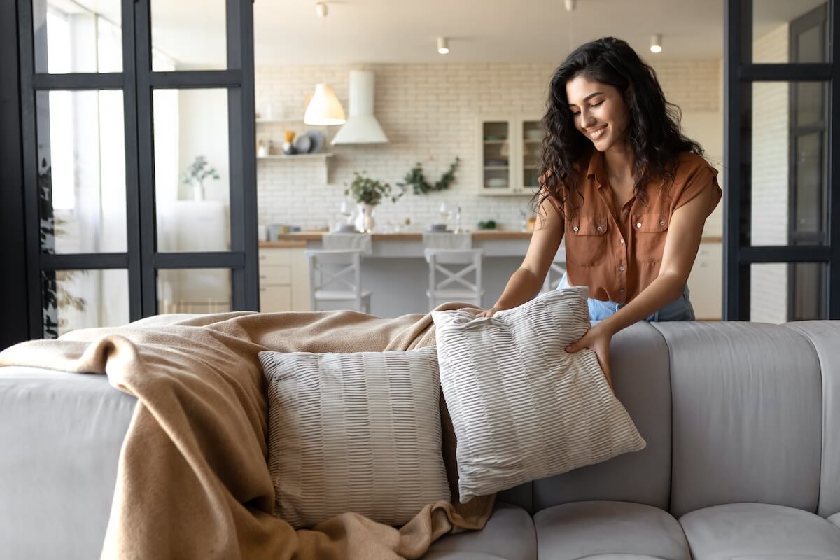 Shutterstock: Lovely young woman putting soft pillows and plaid on comfy sofa, making her home cozy and warm, copy space. Millennial lady decorating her couch with cushions and blanket for autumn or winter