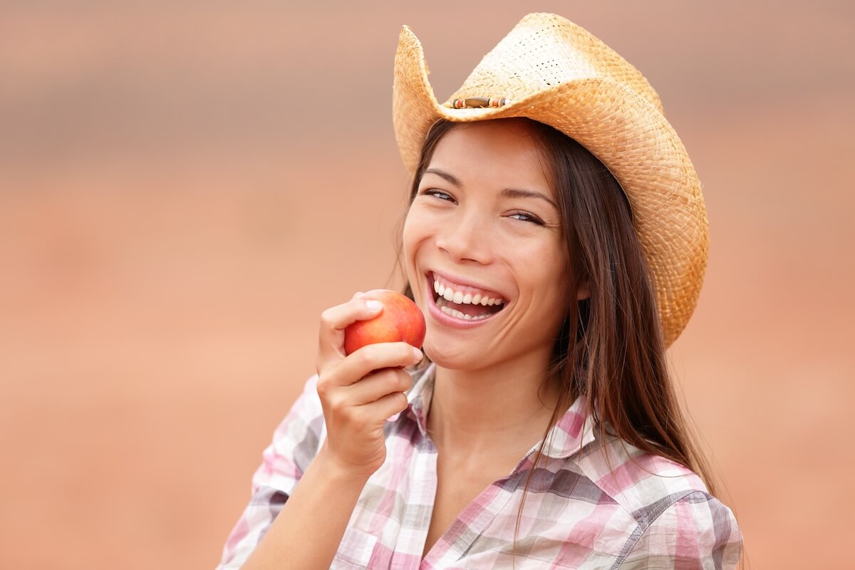 Shutterstock: American cowgirl eating peach or nectarine fruit smiling and laughing happy wearing cowboy hat outside. Healthy eating concept with beautiful young mixed race Caucasian Asian female model outdoor.