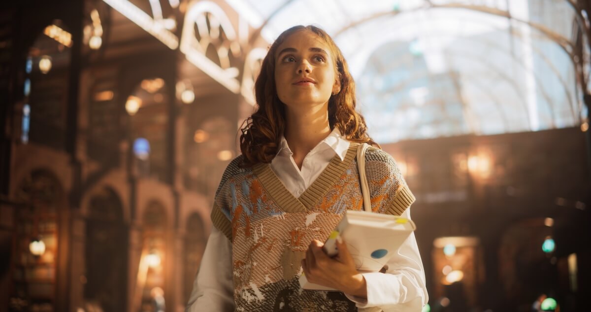 Shutterstock: Young Caucasian Female Student Holding Books in a Bustling Train Station. Intelligent Woman with Curly Hair, Wearing a Sweater Vest and White Shirt, Looking Upward with Hopeful Expression