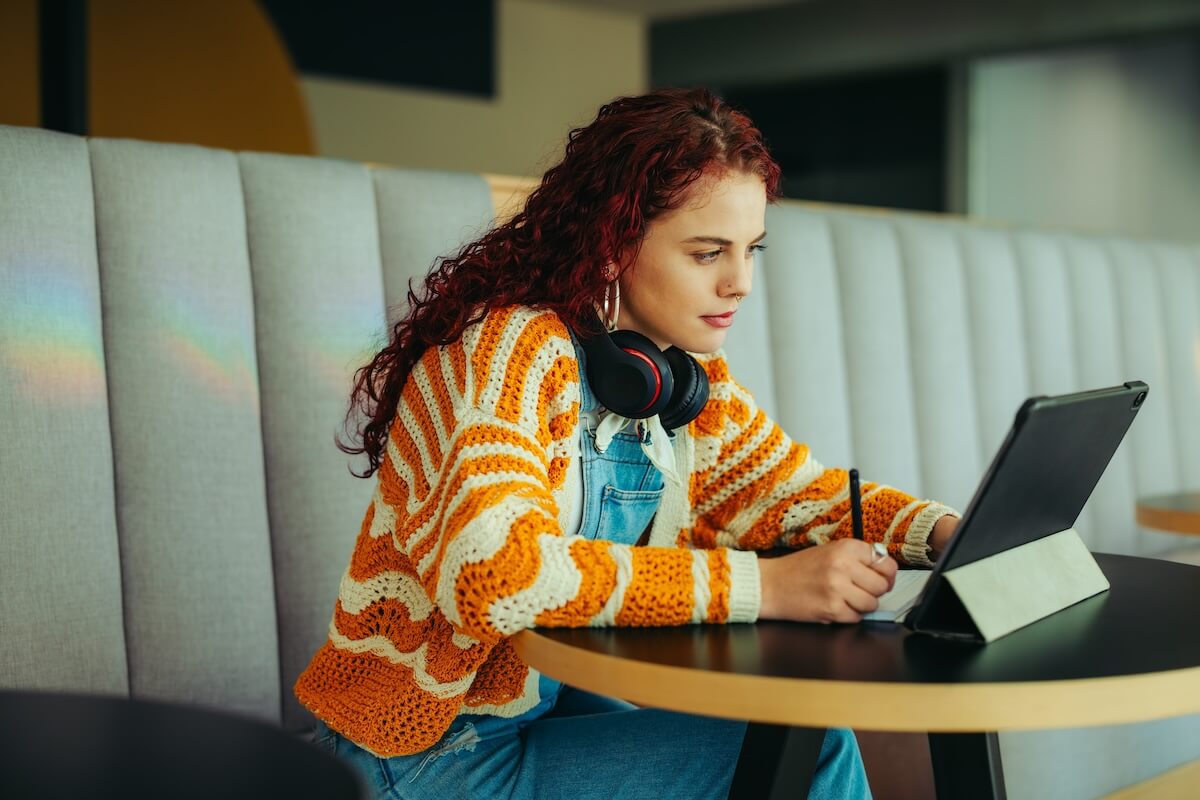 Shutterstock: A female student intently studies with a tablet, wearing headphones and a colorful striped sweater, seated in a cozy, modern cafe environment.