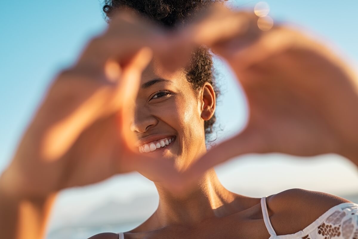 Shutterstock: Close up of hands of young woman making heart shape. Portrait of a lovely black woman making heart with fingers while looking at camera. Smiling african american woman making love sign during sunset.