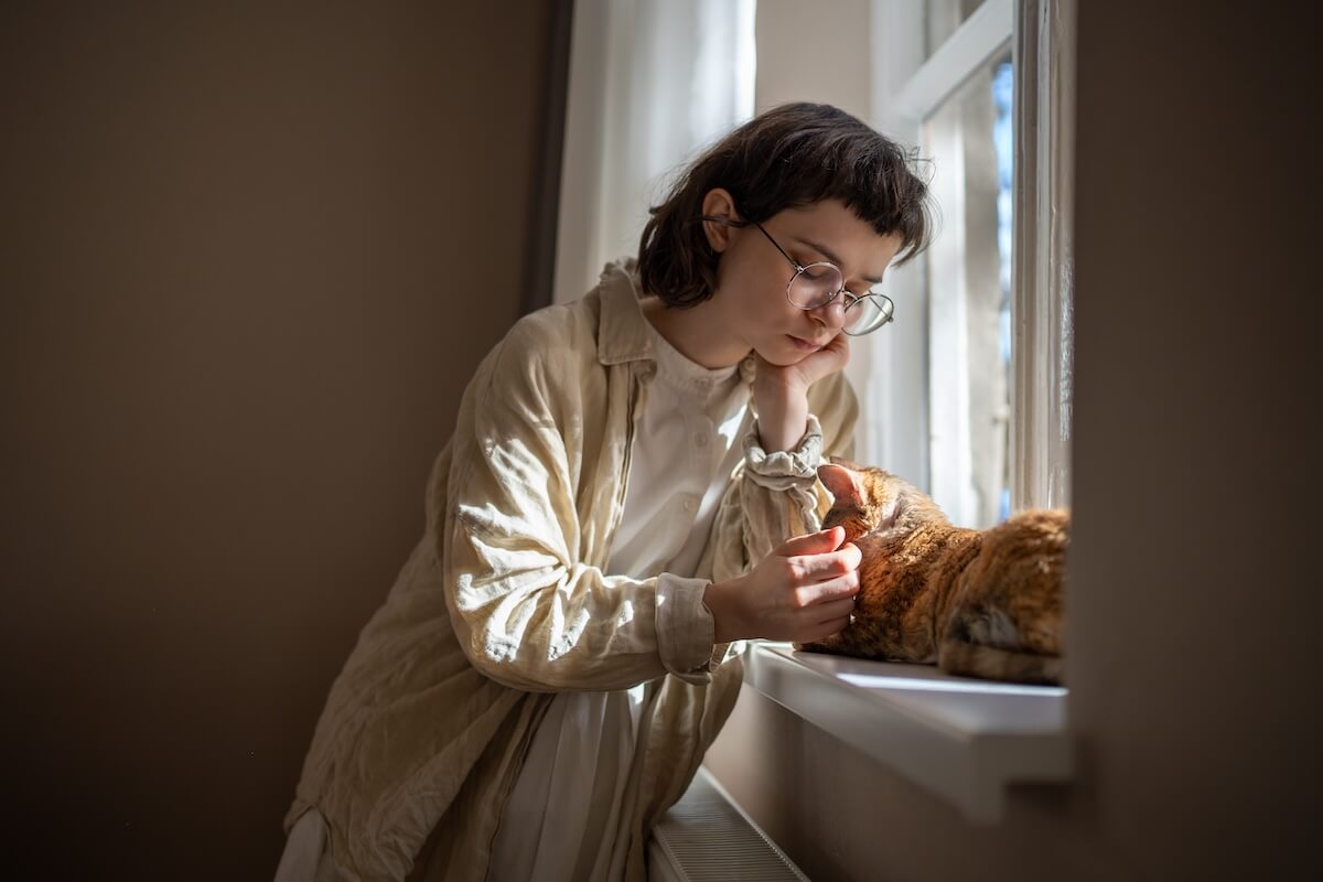 Shutterstock: Lonely teenage girl need of emotional support, tactile contact standing near window with adorable cat friend. Caring young woman stroking cat sadly looking thinking life troubles. Pet therapy at home.