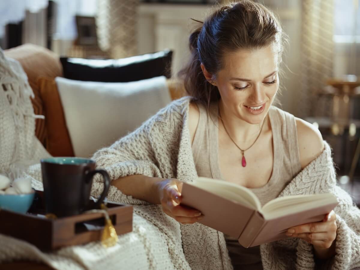 Shutterstock: happy young woman in knitted cosy cardigan with tray and cup reading book at modern home in sunny autumn day.