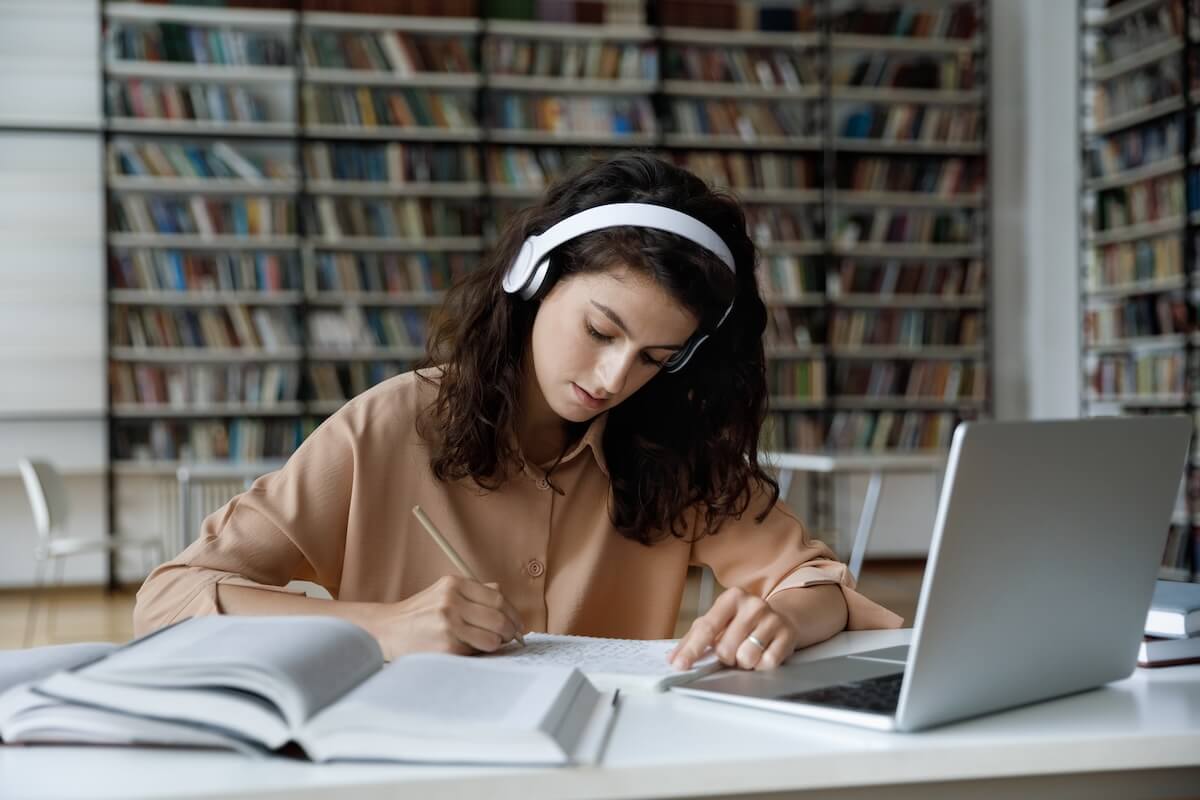 Shutterstock: Serious busy hardworking student girl in headphones working essay, study project in college public library, attending online learning conference, watching video lesson on internet, writing notes