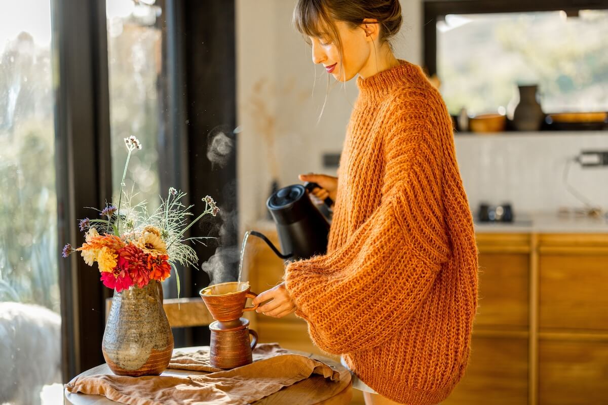 Shutterstock: A woman in an oversized cozy orange sweater prepares pour-over coffee, with warm sunlight filling the rustic kitchen