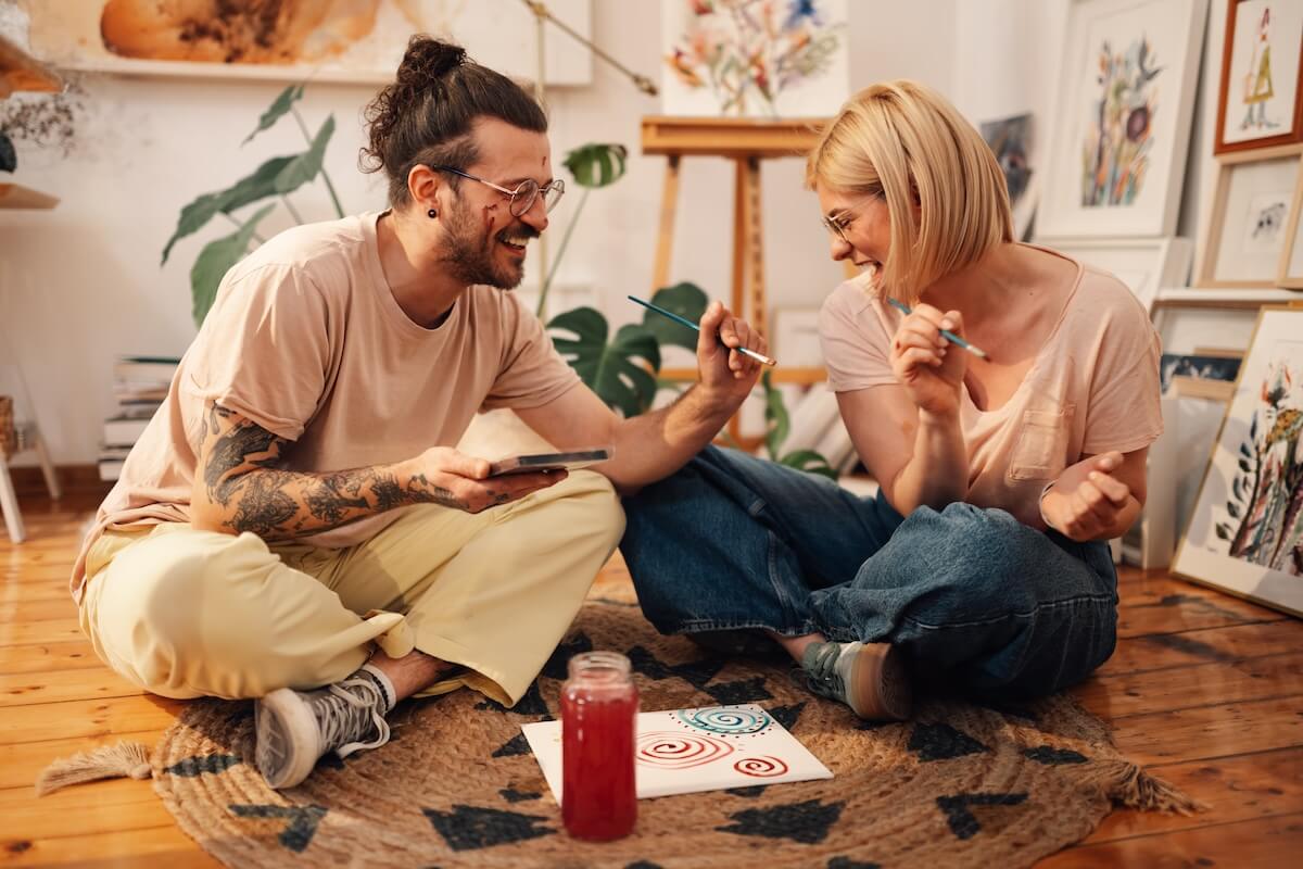 Shutterstock: Smiling artistic fun couple sitting on a floor at creative art studio and acting childish. Young hipster painter couple painting on each other face and having fun together at creative atelier.