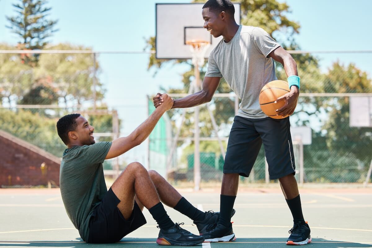 Shutterstock: Basketball, sports and teamwork, helping hand and support, respect and assistance in competition training games. Happy basketball player holding hands with friend, trust and kindness on outdoor court