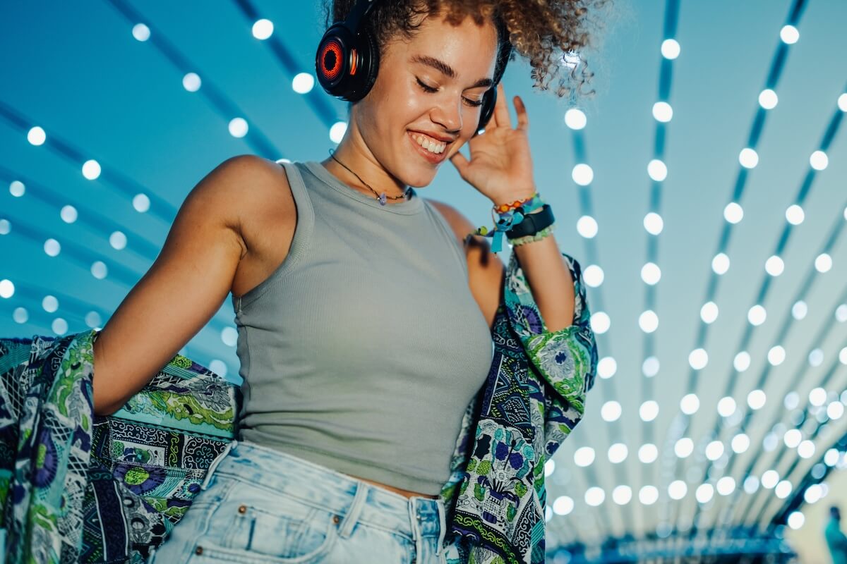 Shutterstock: Happy young woman dancing joyfully at a vibrant music festival, wearing headphones and surrounded by decorative lights, fully embracing the fun and excitement of summer nightlife