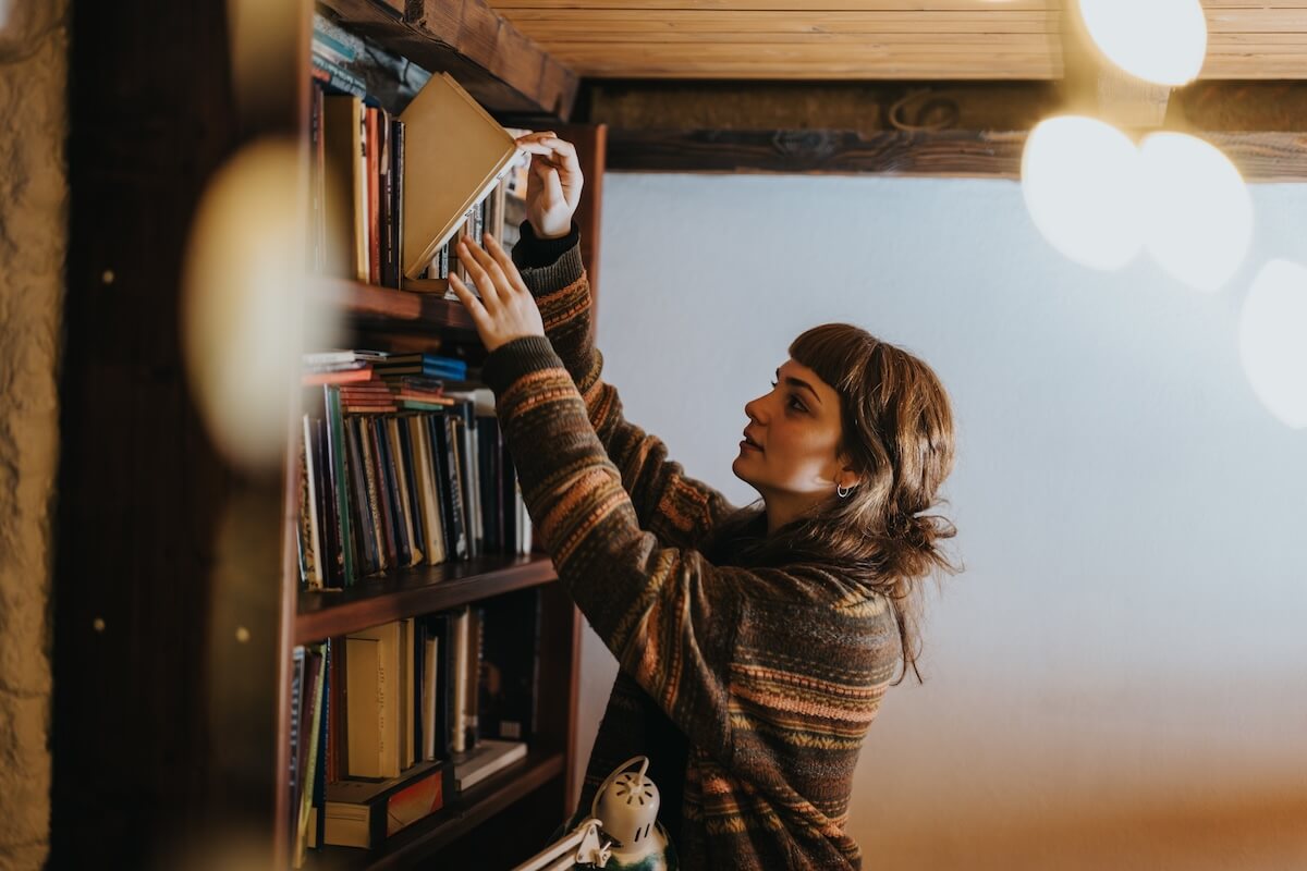Shutterstock: A woman in a warm, rustic setting reaches for a book on a wooden shelf. The ambiance reflects a tranquil and intellectual mood within an inviting home library or study room.