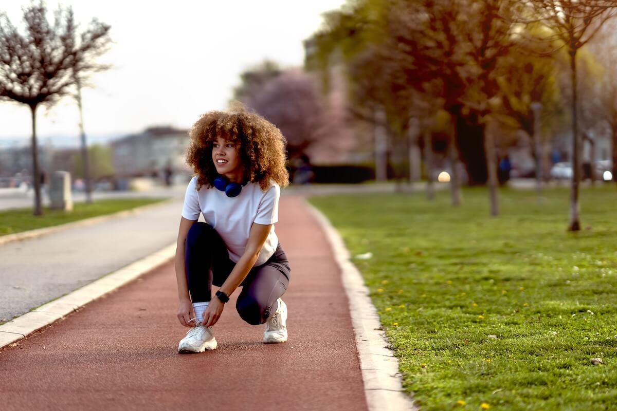 Shutterstock: Young woman tying shoelaces on a running track in an urban park during sunset. She is dressed in sporty attire with headphones around her neck, preparing for a jog.