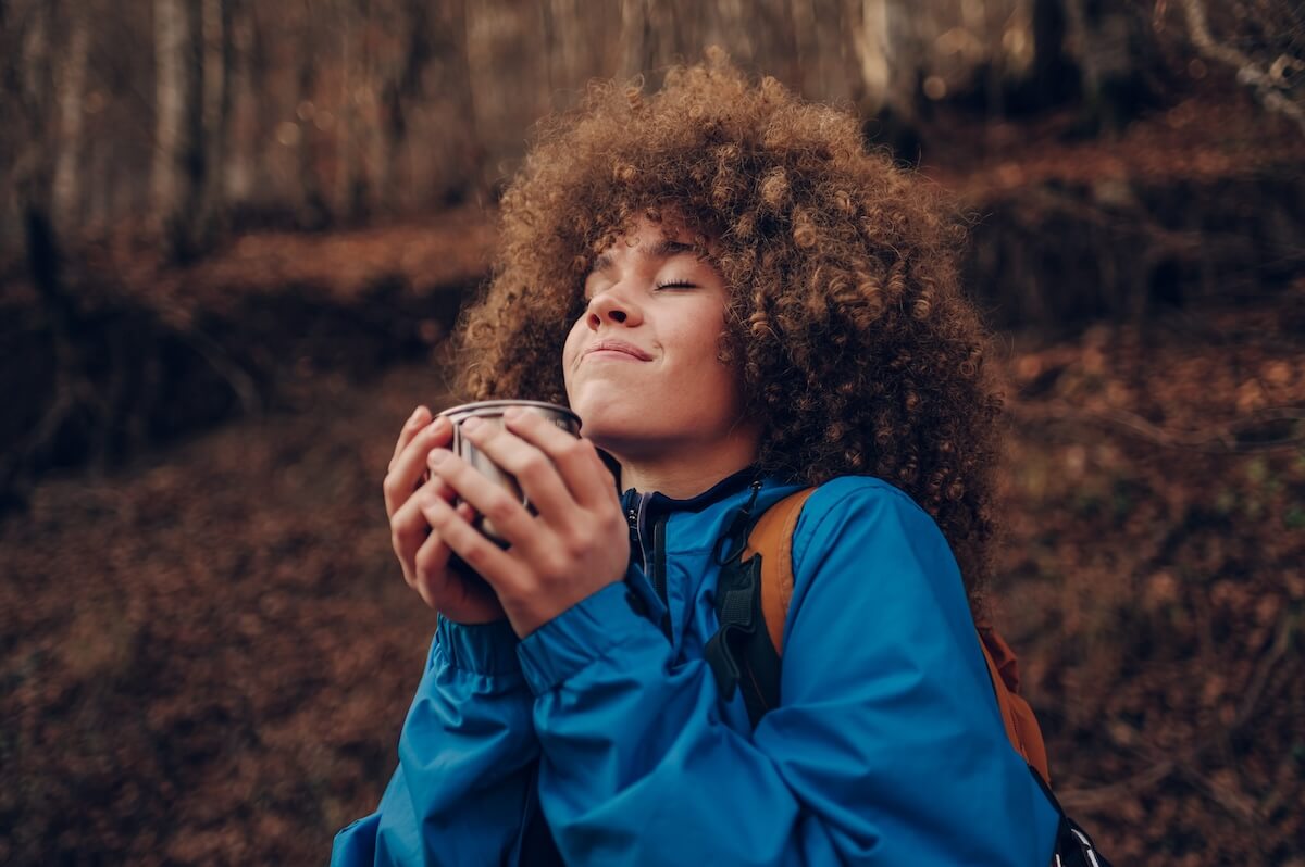 Shutterstock: Hiker sipping from a metal cup, basking in the warmth while surrounded by the vibrant colors of an autumn forest