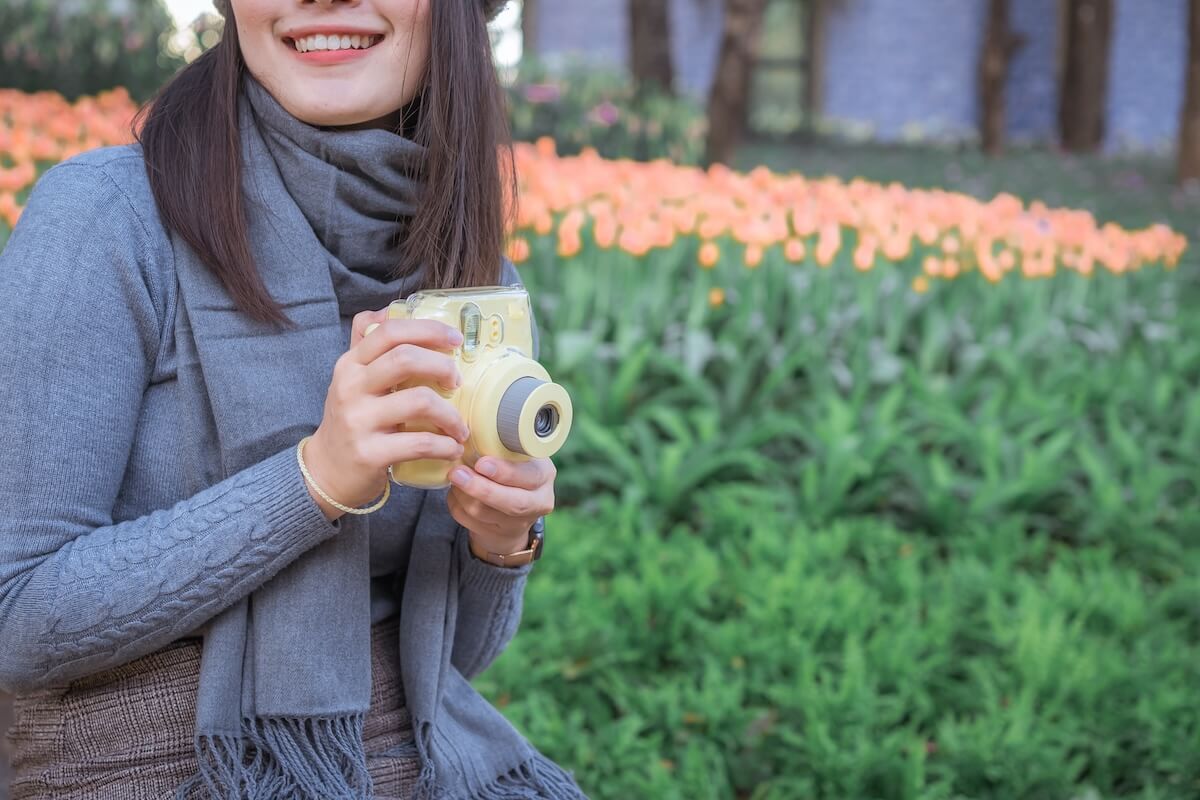 Shutterstock: Happy Asian woman smiling in woolen grey cap holding and using Polaroid camera in garden.